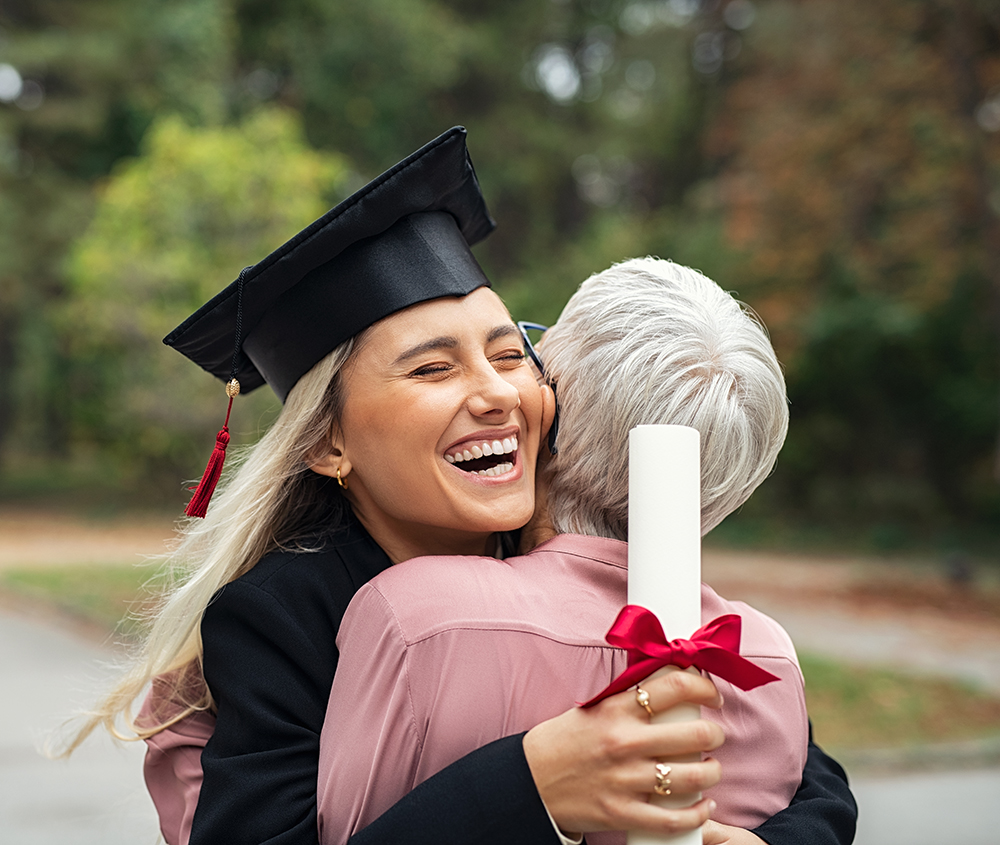 Enthusiastic graduated daughter holding degree embracing mother in campus. Young female student graduate hugging her mother at graduation ceremony. Excited college student with the graduation gown and hat holding diploma and hug the parent at campus.